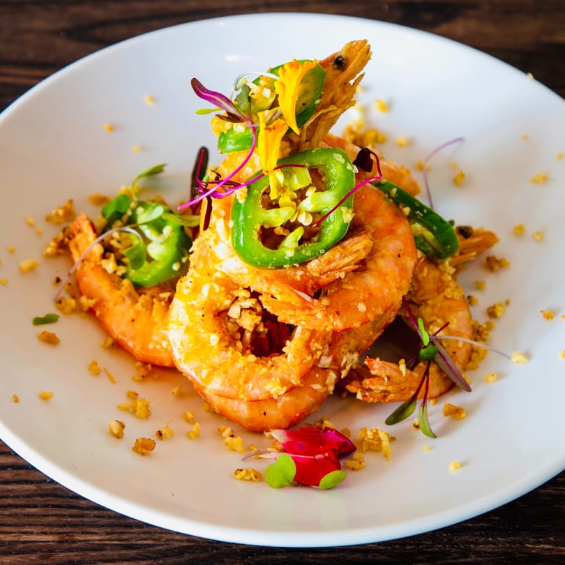 Crispy fried shrimp with sliced green chilies, microgreens, and edible flowers on a white plate.